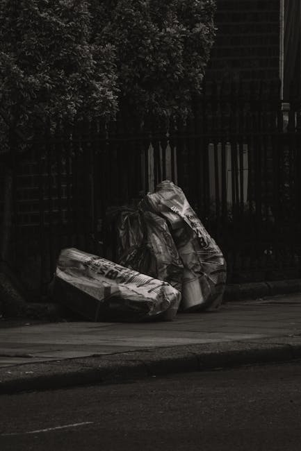 Two large plastic bags of mixed waste material, including cardboard boxes and other refuse, are piled on the pavement beside a black metal fence and a hedge. The bags are crumpled and unevenly stacked